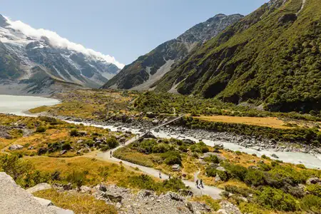 First swing bridge on the Hooker Valley Track — sunshine, silence, and the grandeur of Aoraki. © Anna Jackowska Landscape view of the Hooker Valley Track with the first swing bridge and silhouettes of hikers in the distance on a sunny day in New Zealand’s Southern Alps.