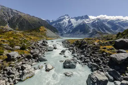 Glacial river flowing through the dramatic landscape of Aoraki/Mount Cook National Park, South Island, New Zealand A wide shot of a milky turquoise glacial river winding through a rocky valley in Aoraki/Mount Cook National Park, with snow-capped mountains and scattered clouds in the background.