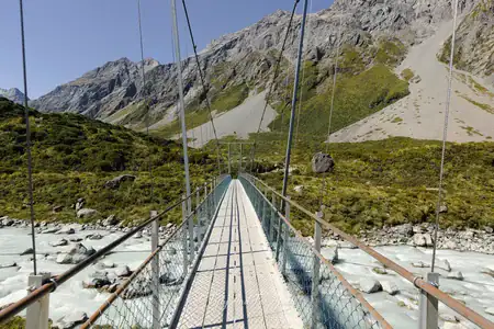 Crossing the suspension bridge over the Hooker River on the Hooker Valley Track. Photo copyright Anna Jackowska. A perspective view from the entrance of a long suspension bridge with a wooden walkway and metal railings, stretching over a milky turquoise river in a rocky valley with steep mountains on either side.