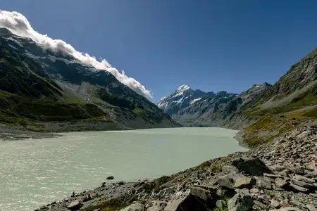 The tranquil waters of Hooker Lake with Mount Cook in the distance. Photo copyright Anna Jackowska. A wide view of Hooker Lake, a milky turquoise glacial lake, surrounded by rocky terrain and towering mountains, with the snow-capped Mount Cook visible in the center against a blue sky with some clouds.