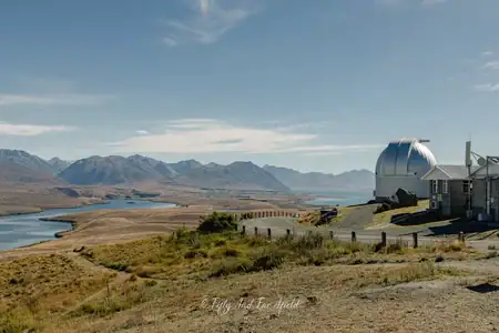 Mount John University Observatory with a panoramic view of Lake Tekapo and the Southern Alps. A wide view of the Mount John University Observatory with its silver dome on a hilltop overlooking the turquoise waters of Lake Tekapo and the distant Southern Alps under a partly cloudy blue sky.