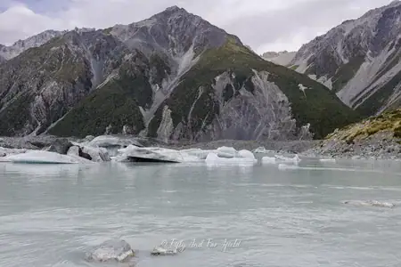 Icebergs floating on the milky waters of Tasman Lake, Aoraki/Mount Cook National Park. A view of Tasman Lake with several white and grey icebergs floating on its milky turquoise surface, set against a backdrop of tall, rugged mountains under a cloudy sky in Aoraki/Mount Cook National Park.