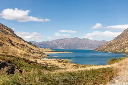 Panoramic view of Lake Hāwea from a scenic lookout. A wide panoramic view of the deep blue waters of Lake Hāwea surrounded by dry, golden-brown hills and mountains under a bright blue sky with scattered clouds, as seen from a lookout point.