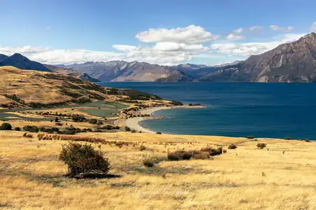 Scenic view of Lake Wanaka and surrounding mountains from a lookout point. A wide panoramic view of Lake Wanaka's deep blue waters and distant mountain ranges under a partly cloudy sky, as seen from a lookout point with golden-brown hills and a curved shoreline in the foreground.