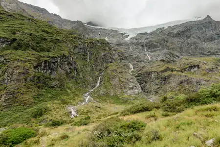 View of Rob Roy Glacier from the track and lookout, Mt Aspiring National Park, New Zealand. Photo copyright Anna Jackowska. A view of a glacier situated high on rugged mountains with multiple waterfalls flowing down, as seen from the Rob Roy's Glacier Track and lookout in Mt Aspiring National Park, under an overcast sky.