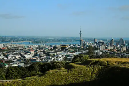 Panoramic view of Auckland city, including the Sky Tower and Harbour Bridge. A wide view of Auckland city from a hilltop, showing the Sky Tower, numerous buildings, the Waitematā Harbour, and the Auckland Harbour Bridge in the distance under a clear blue sky.