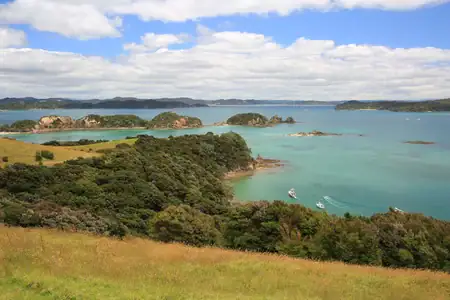 A stunning panoramic vista of the Bay of Islands, New Zealand. A wide panoramic view of the Bay of Islands with numerous green islands, turquoise water with white boats, and a grassy hillside in the foreground under a blue sky with white clouds.