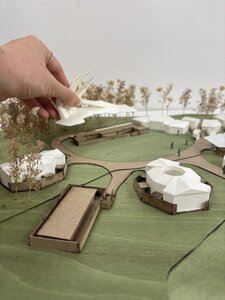 Architectural site model of campus pavilions with oculus, curving paths, trees, and designer’s hand.