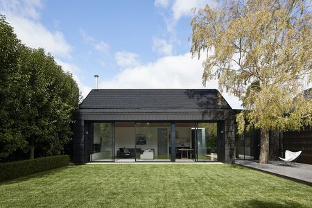 Modern single-story house with glass facade, manicured lawn, hedge, tree, and patio area.
