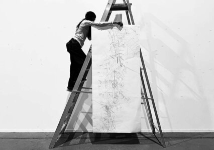 Black-and-white studio: artist on ladder drawing intricate lines on long paper scroll.
