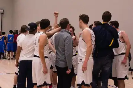 A basketball team in white uniforms huddles together on the court, raising hands in a gesture of unity before a game at Telos Academy. Players in blue are visible in the background. at Telos
