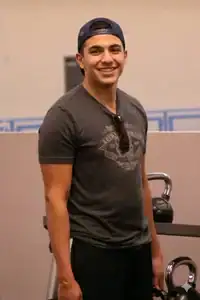Young man in a dark t-shirt and backward cap stands in a gym, smiling, with a kettlebell and gym equipment visible in the background—part of the supportive environment at Telos U, where tailored outpatient treatment helps individuals thrive. at Telos