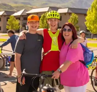 A mother, a son, and another son who is a Telos student pose together outdoors at a biking event, with one person in cycling gear and helmet, and two others—smiling by their side—representing Telos Academy. Buildings and trees are visible in the background