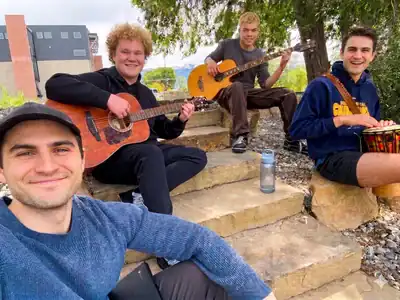 Four young men sit outdoors on stone steps at Telos U; two play guitars, one plays a drum, and one smiles at the camera. A water bottle sits nearby, and buildings and trees are in the background—capturing a moment from their Boys Residential Treatment experience. at Telos