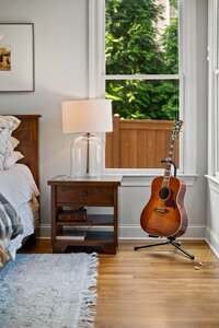 Bright bedroom featuring a guitar, wooden bedside table, and large window with lush greenery outside.
