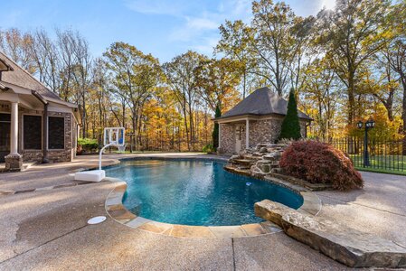 Bright backyard pool area surrounded by trees with autumn foliage, featuring waterfall and cozy poolside ambiance.