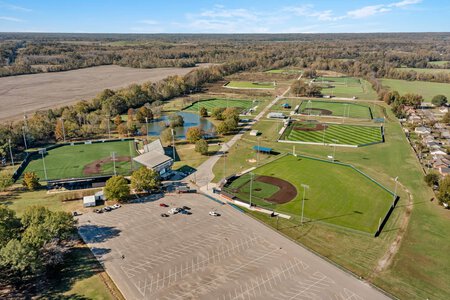Aerial view of a baseball and softball complex with multiple fields, parking lot, and surrounding landscape under blue sky.