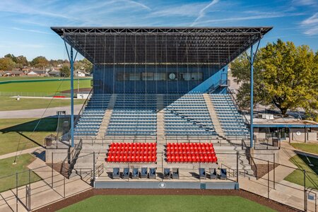 Brightly lit stadium with blue seats and red chairs under a large canopy.