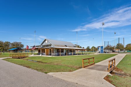 Bright, clear day view of a sports clubhouse with picnic tables, flags, and sports fields in the background.
