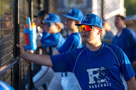 Young baseball player in uniform and sunglasses at dugout.