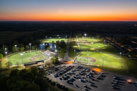 Aerial view of illuminated baseball fields at night with parking area and sunset in background.