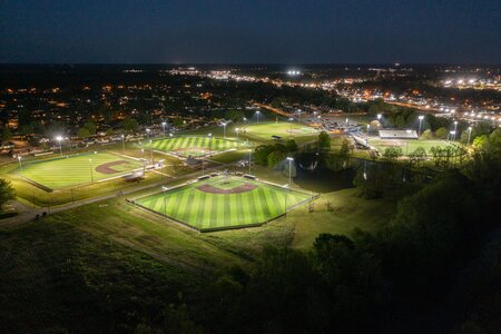 Nighttime aerial view of lit baseball fields in a city park with surrounding neighborhood lights.