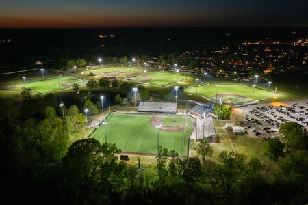 Night view of well-lit baseball complex with multiple fields and parking lot.