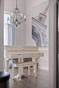 Charming white grand piano with matching bench, illuminated by a crystal chandelier, interior decor with Parisian Eiffel Tower artwork.
