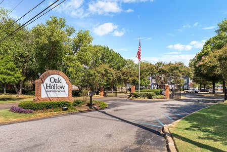 Gated Oak Hollow apartment entrance with American flag, trees, and landscaped surroundings for comfortable living.