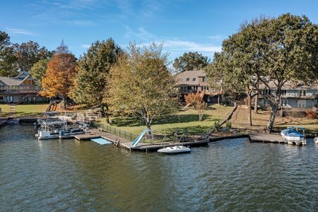 ID: Waterfront home with boat slips on a calm lake, surrounded by mature trees and residential homes in fall colors.