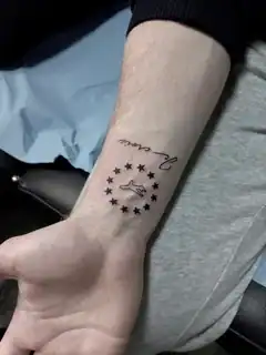 Tatuajes 17 Diseño minimalista en tinta negra con un motivo central rodeado por un círculo de estrellas y una inscripción cursiva, ideal para tatuajes pequeños y delicados.
