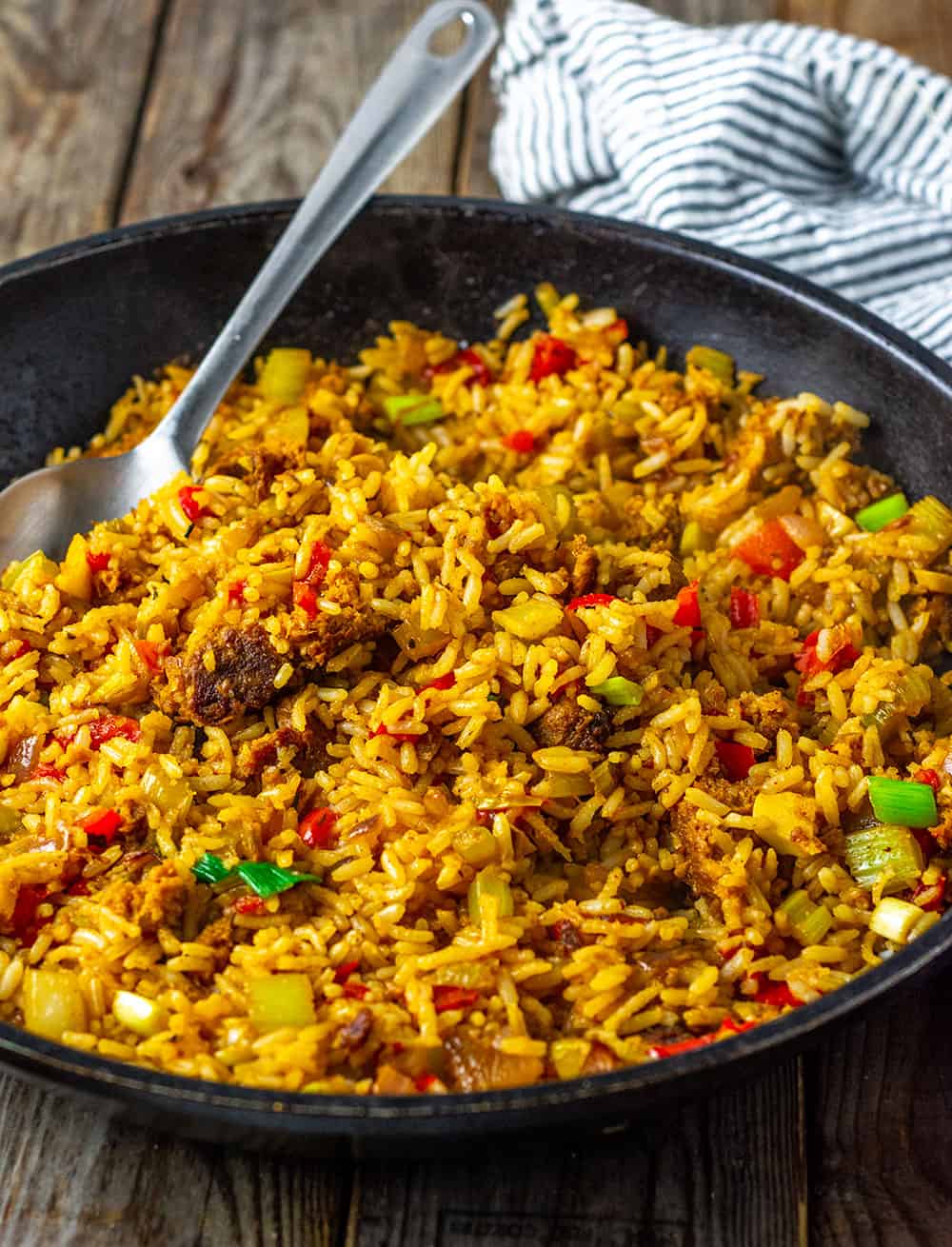 vegan dirty rice, cajun style. Brown rice cooked with onion, garlic, celery and bell peppers with ground burger and Creole seasoning, garnished with green onion in a large black skillet with a silver spoon on a wooden background with a black and white stripe napkin. photo is side view.