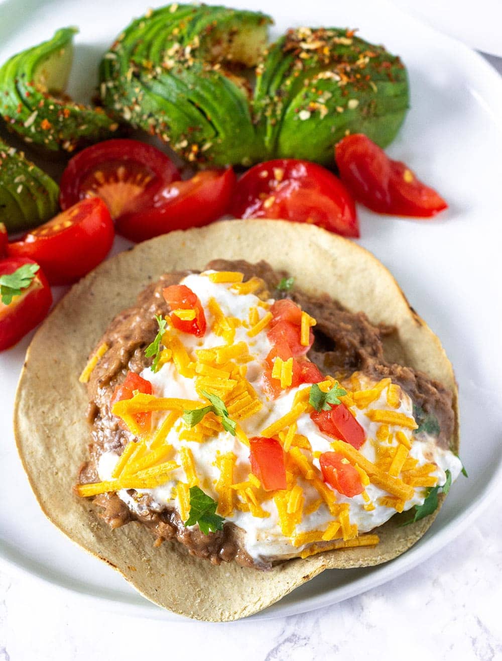 vegan refried beans on tostada garnished with vegan sour cream, tomatoes, vegan cheese with slices of avocados and tomatoes on a white background
