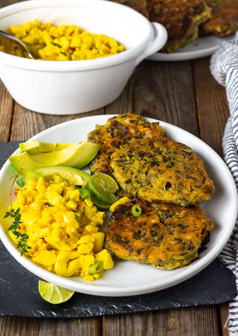 Callaloo Fritters on a white plat with ackee and avocado slices on a wooden background with a white bowl of ackee in the background