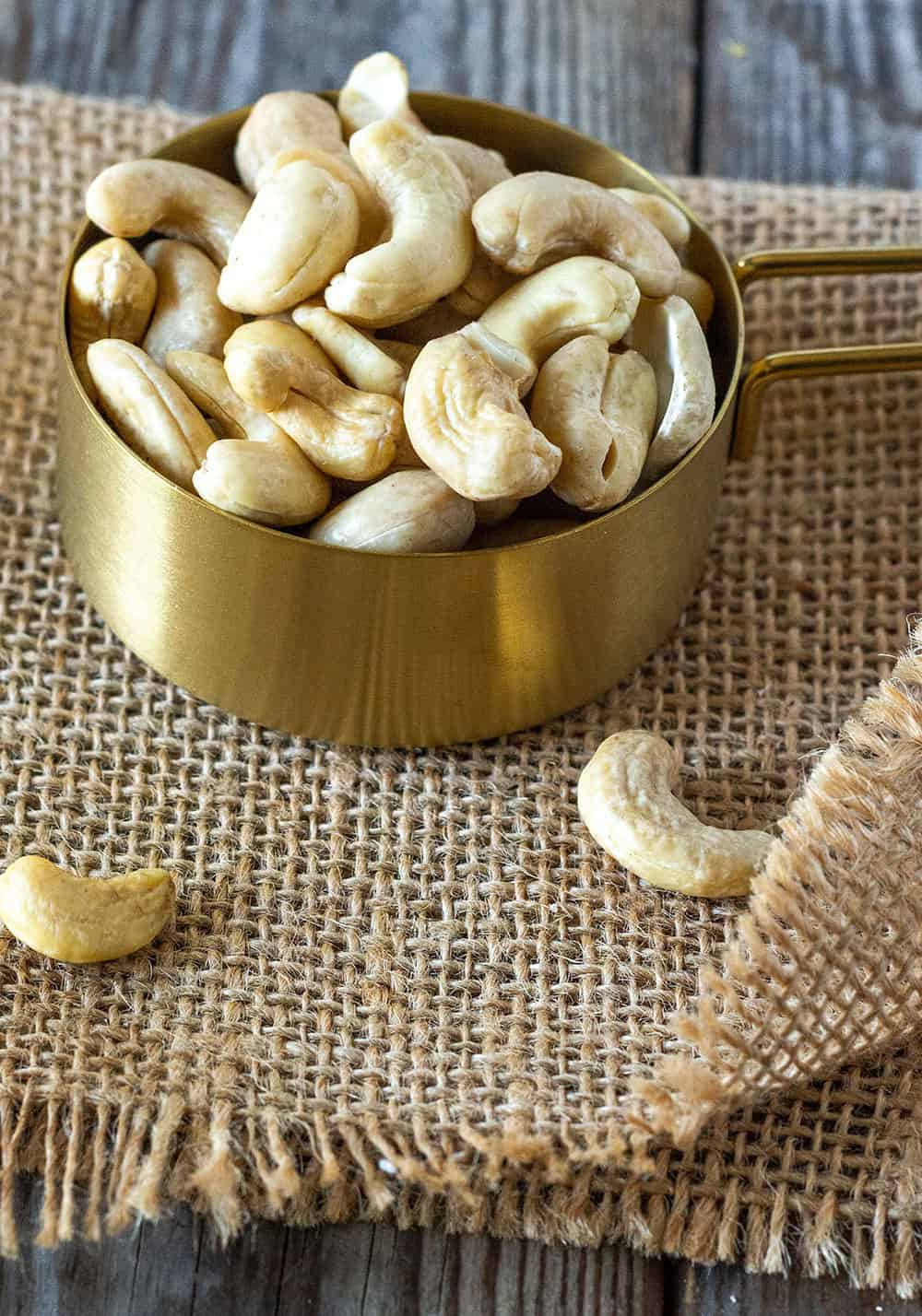 Whole cashews for cashew cream recipe in a gold measuring cup on a brown burlap napkin on a wooden board