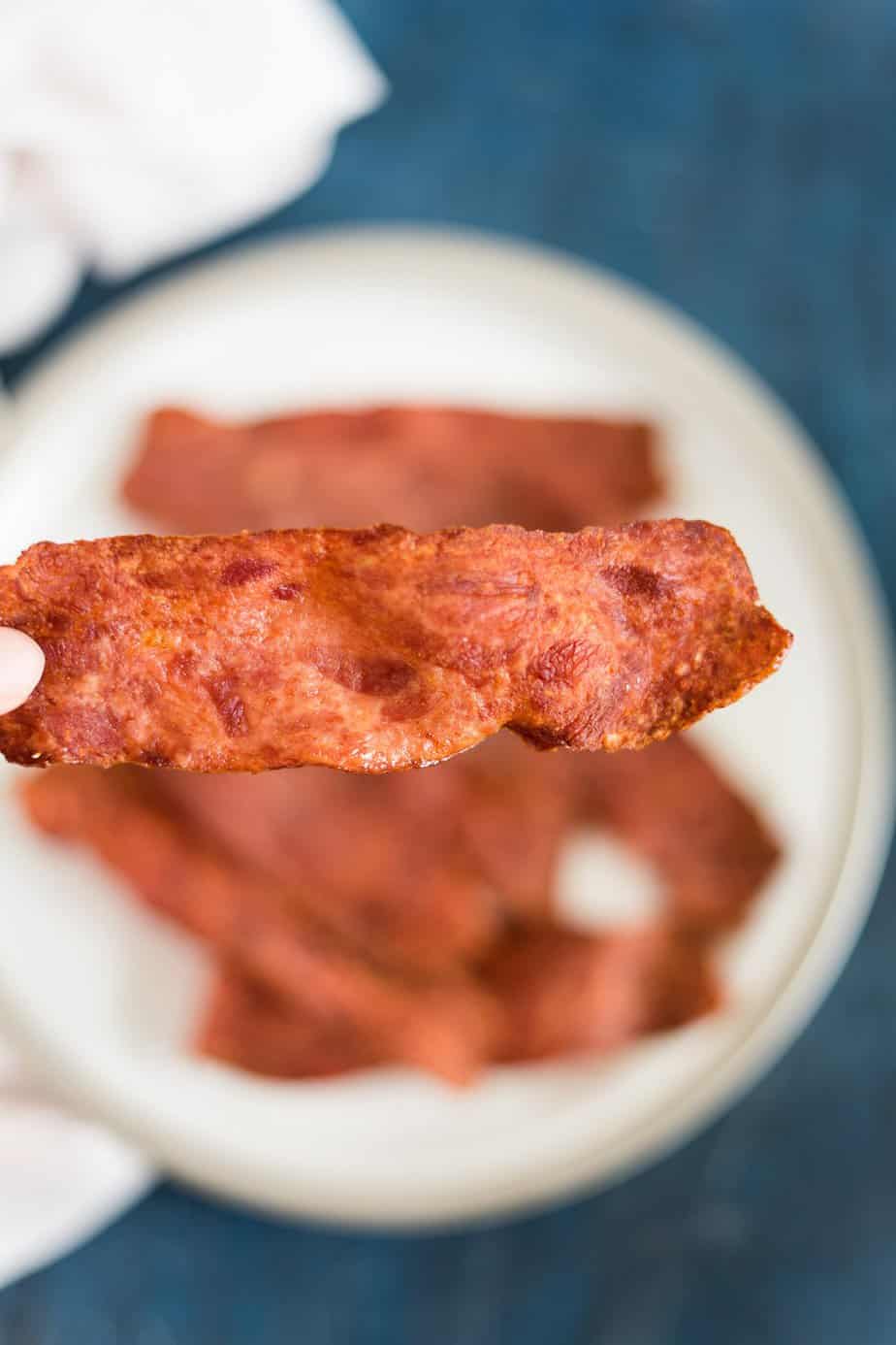 Holding a piece of turkey bacon cooked in the air fryer. behind there is the plate with the other slices.