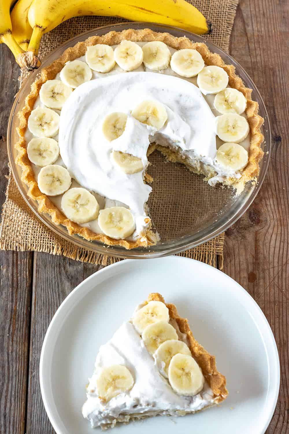 Overlay Banana cream pie in pie plat topped with coconut whipped cream and fresh banana slices with a slice in a white plate on a wooden board