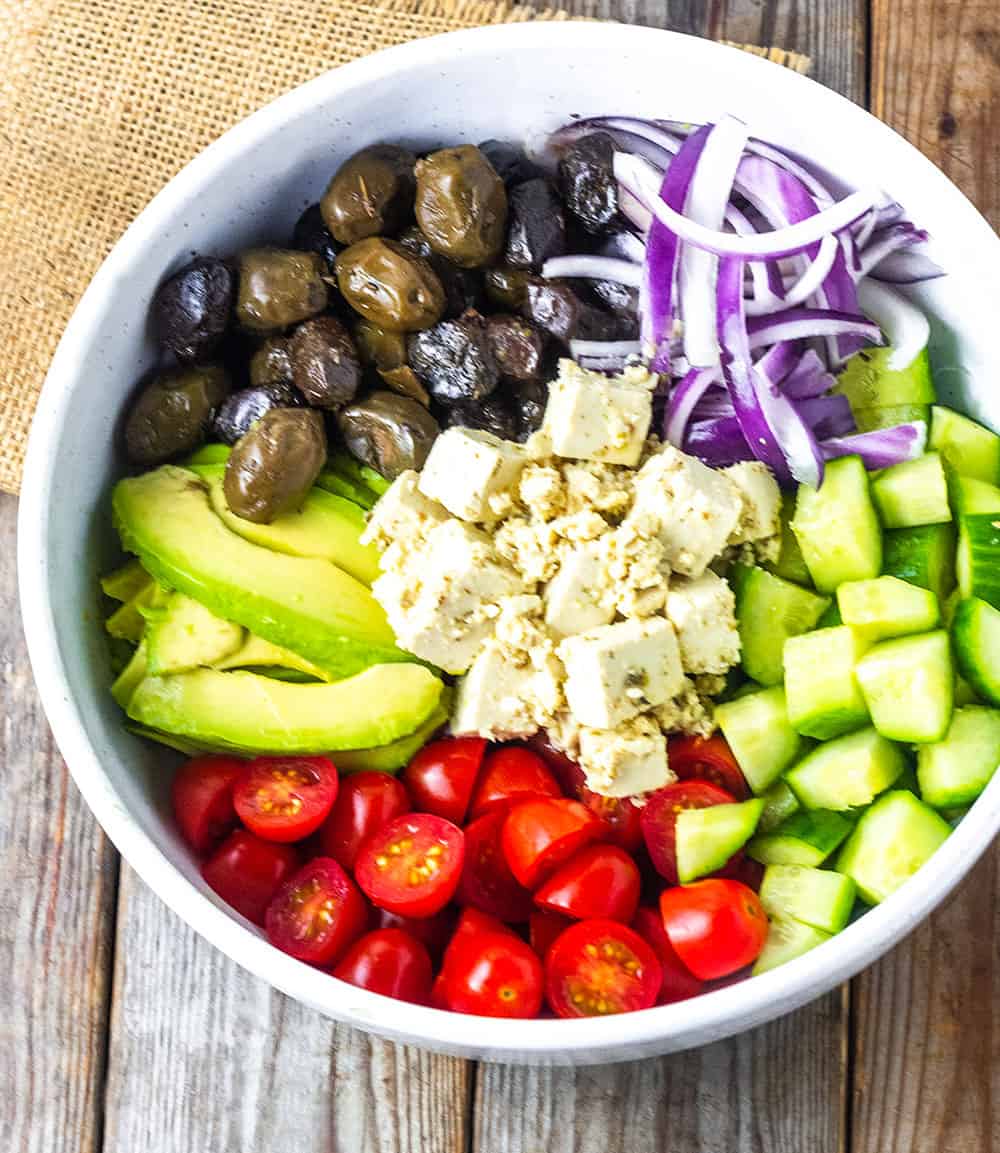 vegan feta cheese in the center of greek salad with chopped tomatoes, cucumber, red onion, olive and avocado in a white bowl on a brown wooden board