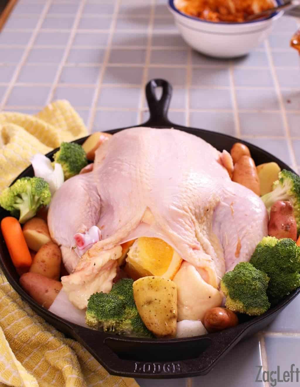 Raw stuffed chicken surrounded by baby carrots, broccoli florets, and potatoes in a cast iron skillet