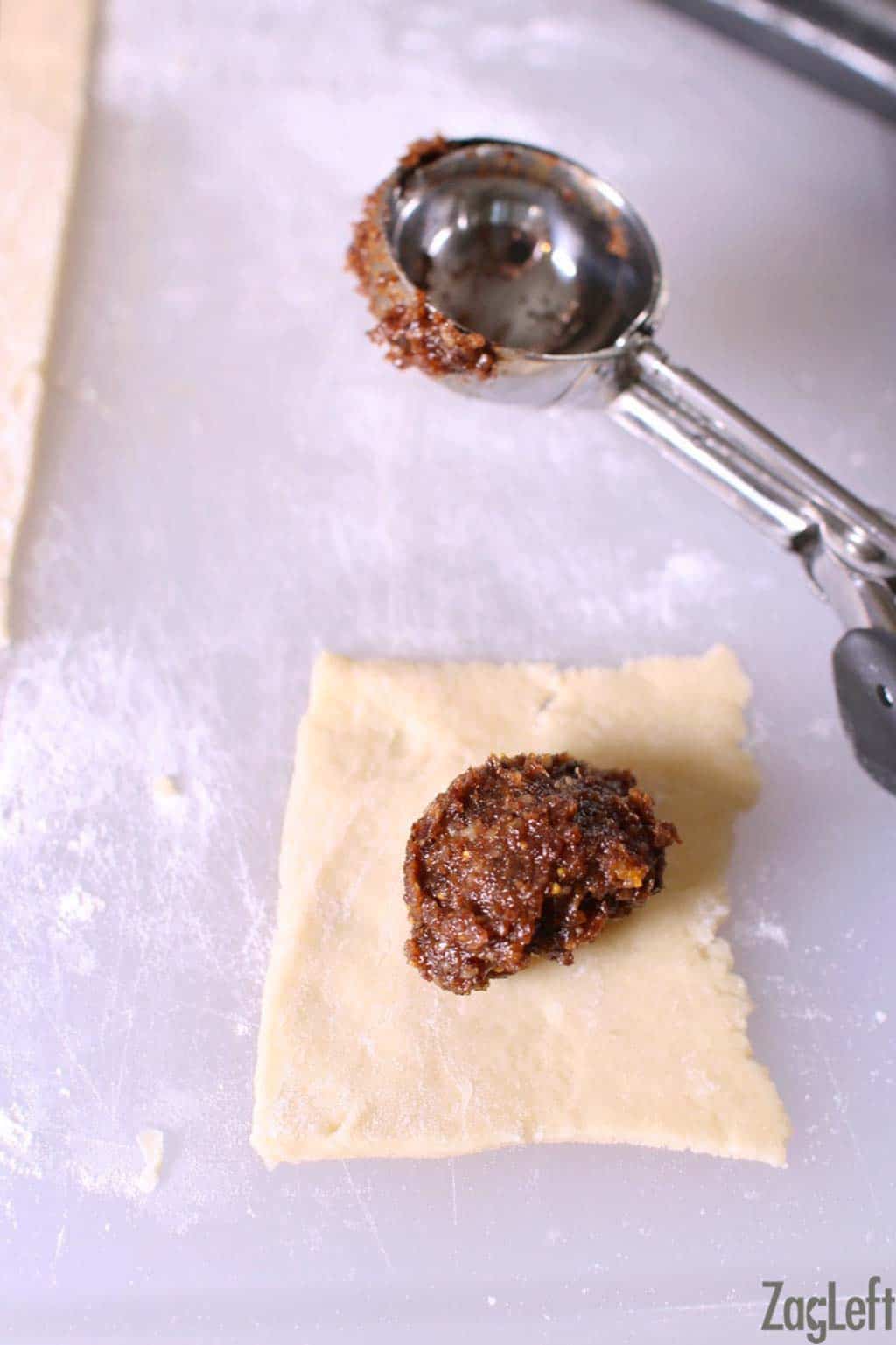 A small rectangle of cuccidati dough topped with a dollop of filling from a cookie scoop on a cutting board