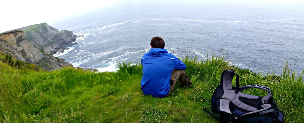 Young man in blue wind coat sits on green grass on edge of cliff overlooking the crashing sea below. It depicts the New Year’s resolutions for the Traveler: take time to wonder.