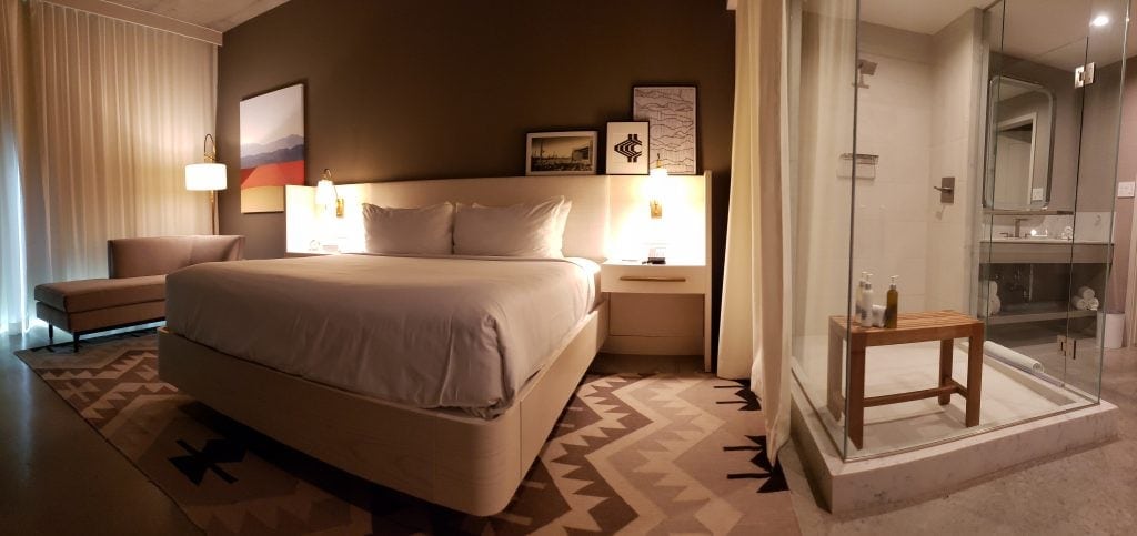 panoramic view of guset room with glass shower enclosure, white bedding, kilim rug in natural tones