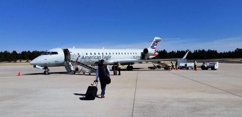 small communter jet sits on Flagstaff travel runway while passenger walks on tarmac with carry on bag - clear skies overhead, trees on other side of runway at airport in Flagstaff Arizona