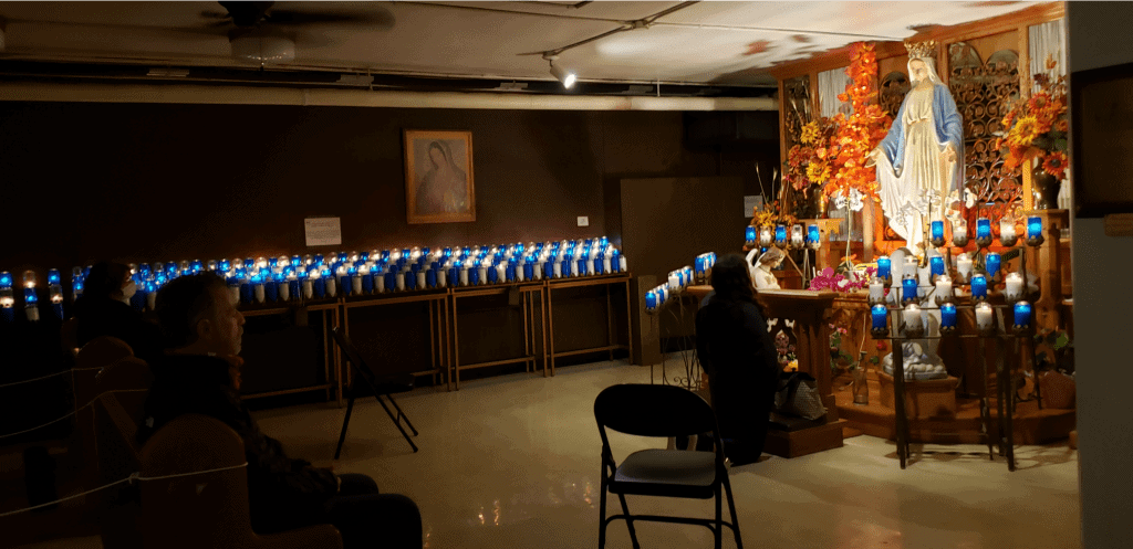Dark, low ceiling room is lined with candles, some in blue glass while people pray in front of a statue of Mary