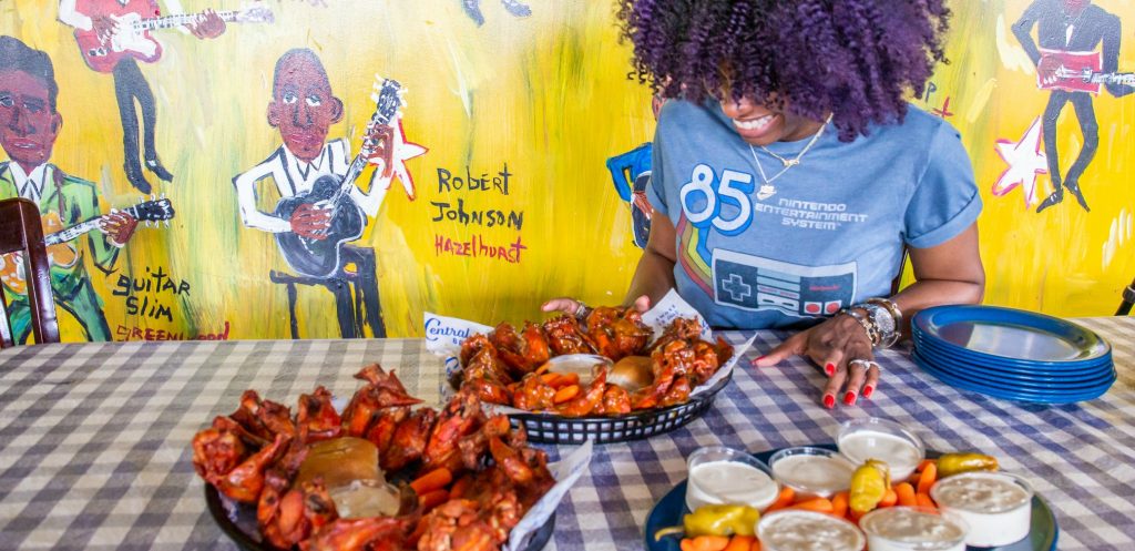 woman looks down at two platters of hot wings - wall of colorful primitive painting behind