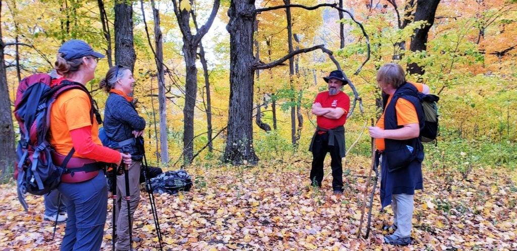 three pilgrims stand facing man in red shirt and black hat - surrounded my trees in fall colors - leaves on ground