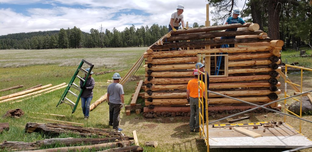 Volunteers in construction helmets work on small Brolliar log cabin