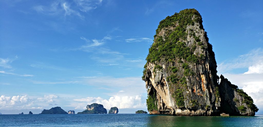 This tall pillar of limestone rock looks more like a sea stack than an island standing high out of green-blue waters. A boat sails in front of the large vertical rock.
