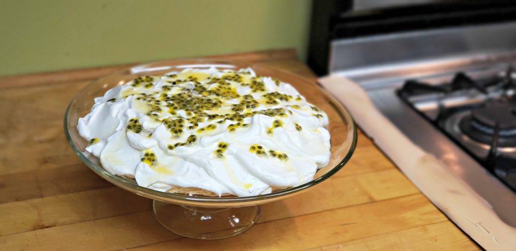 Round passion fruit Pavlova on glass cake holder sits on wooden counter - white whipped cream is dotted with green passion fruit sauce and seeds - just as it comes out of the fruit!