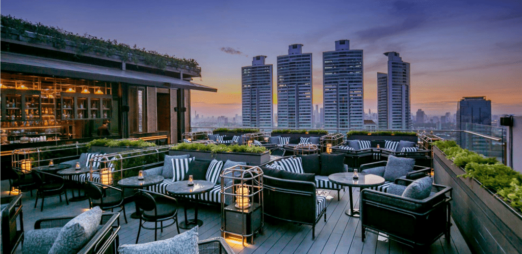 smallish patio bar on the rooftop of Bangkok-outdoor comfy chaise loungs and chairs with views of the highrise BAngkok behind making it a best rooftop bar in Bangkok